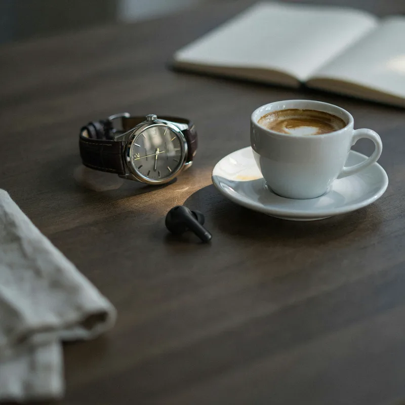 Armbanduhr, Espresso und ein Earbud auf einem dunklen Schreibtisch